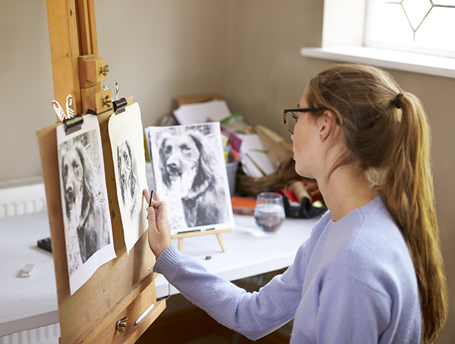 Side View Of Female Teenage Artist Sitting At Easel Drawing Picture Of Dog From Photograph In Charcoal